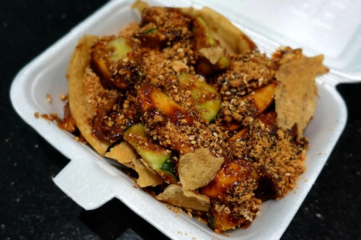 Takeaway box of rojak with cucumber, fruit and fritters under crushed peanuts and sesame