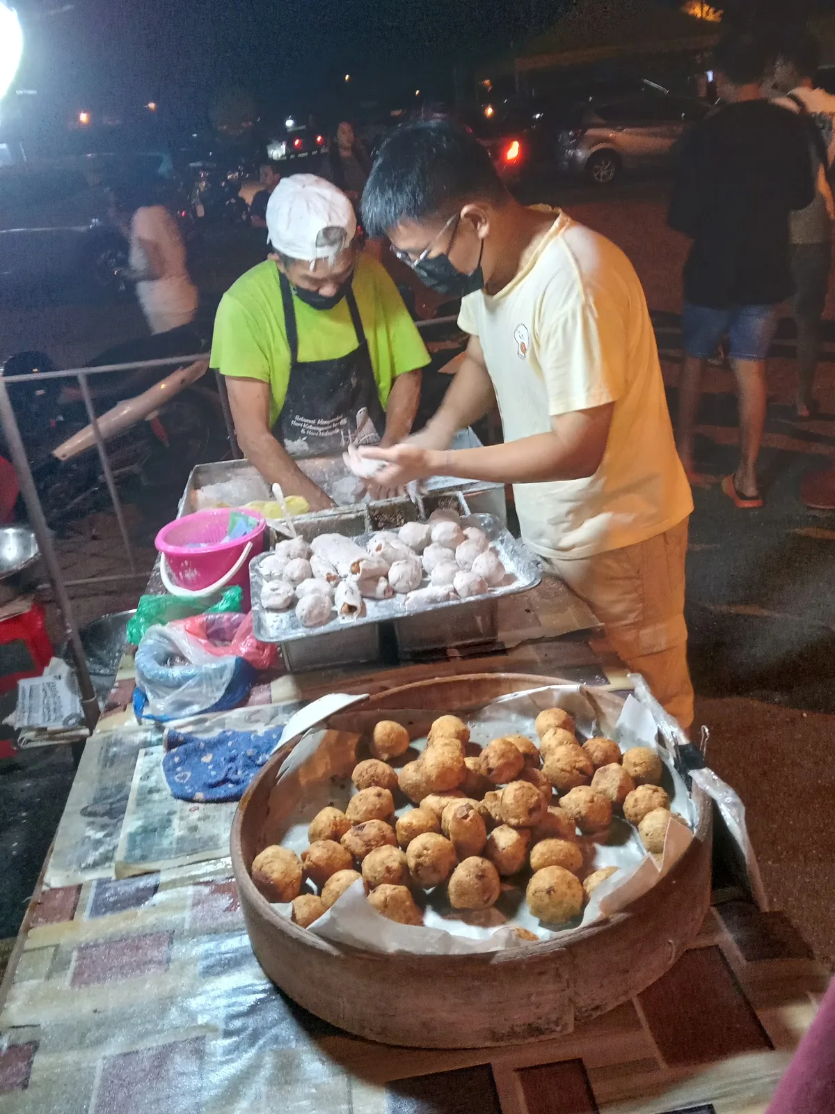 Night market stall with two vendors, steamer trays of buns and a wooden tray of fried dough balls