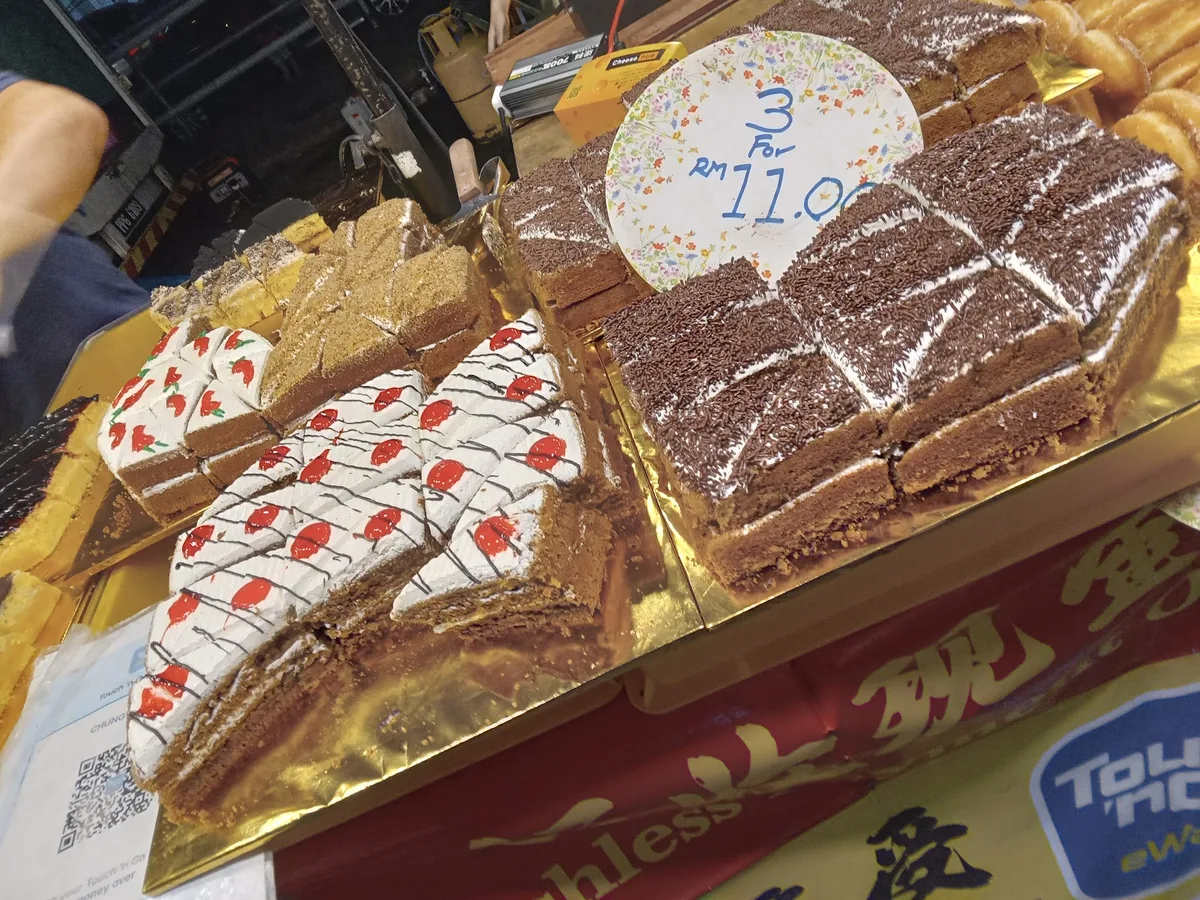 Night market cake stall, sliced sponge cakes on gold trays with a 3 for RM11 sign