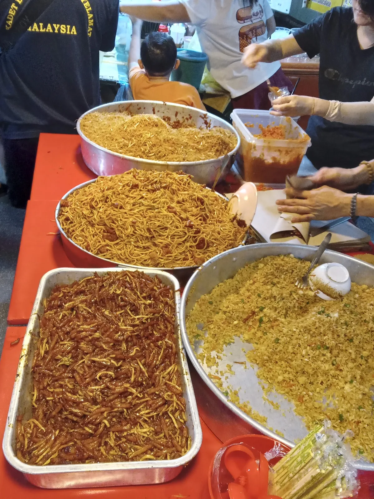 Large trays of fried noodles on a red-clothed stall table with vendors serving