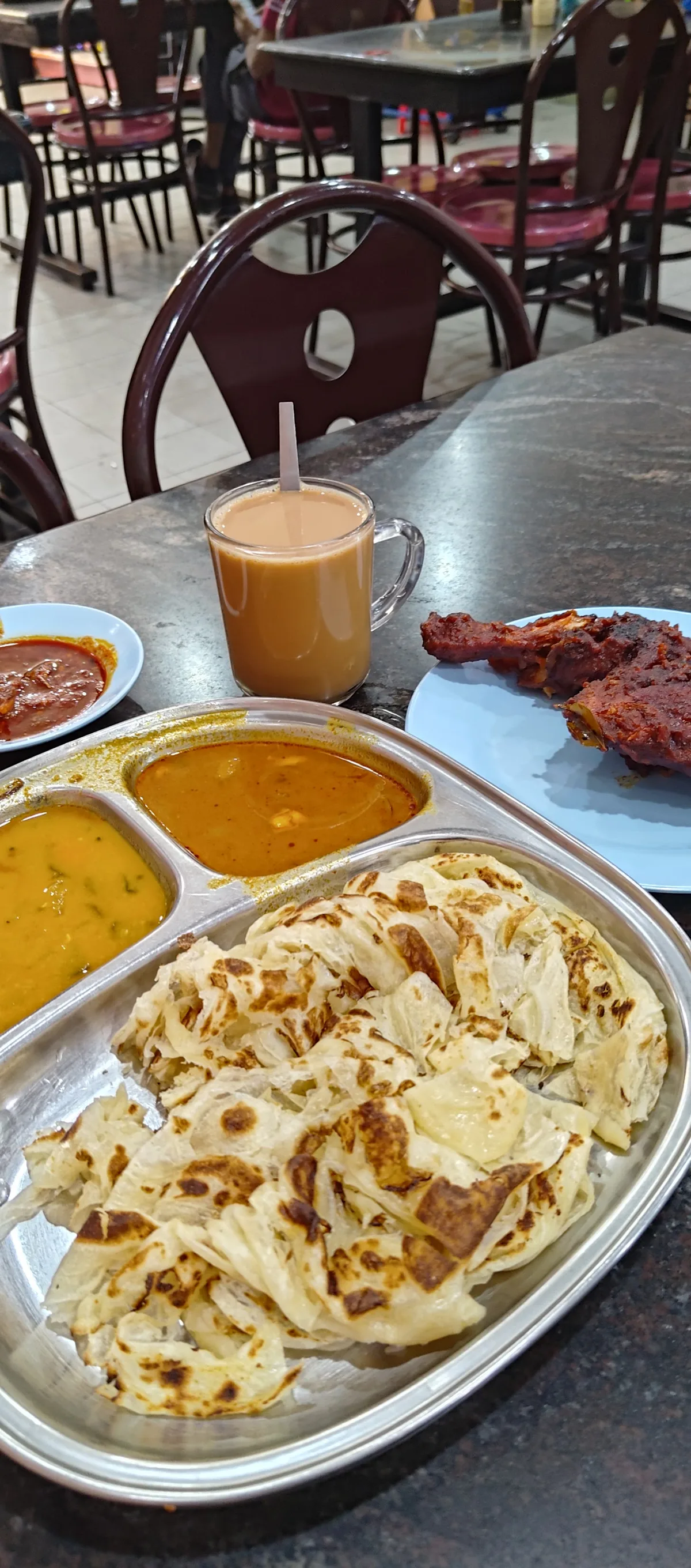 A steel tray of roti canai with two curries, fried chicken and a cup of tea