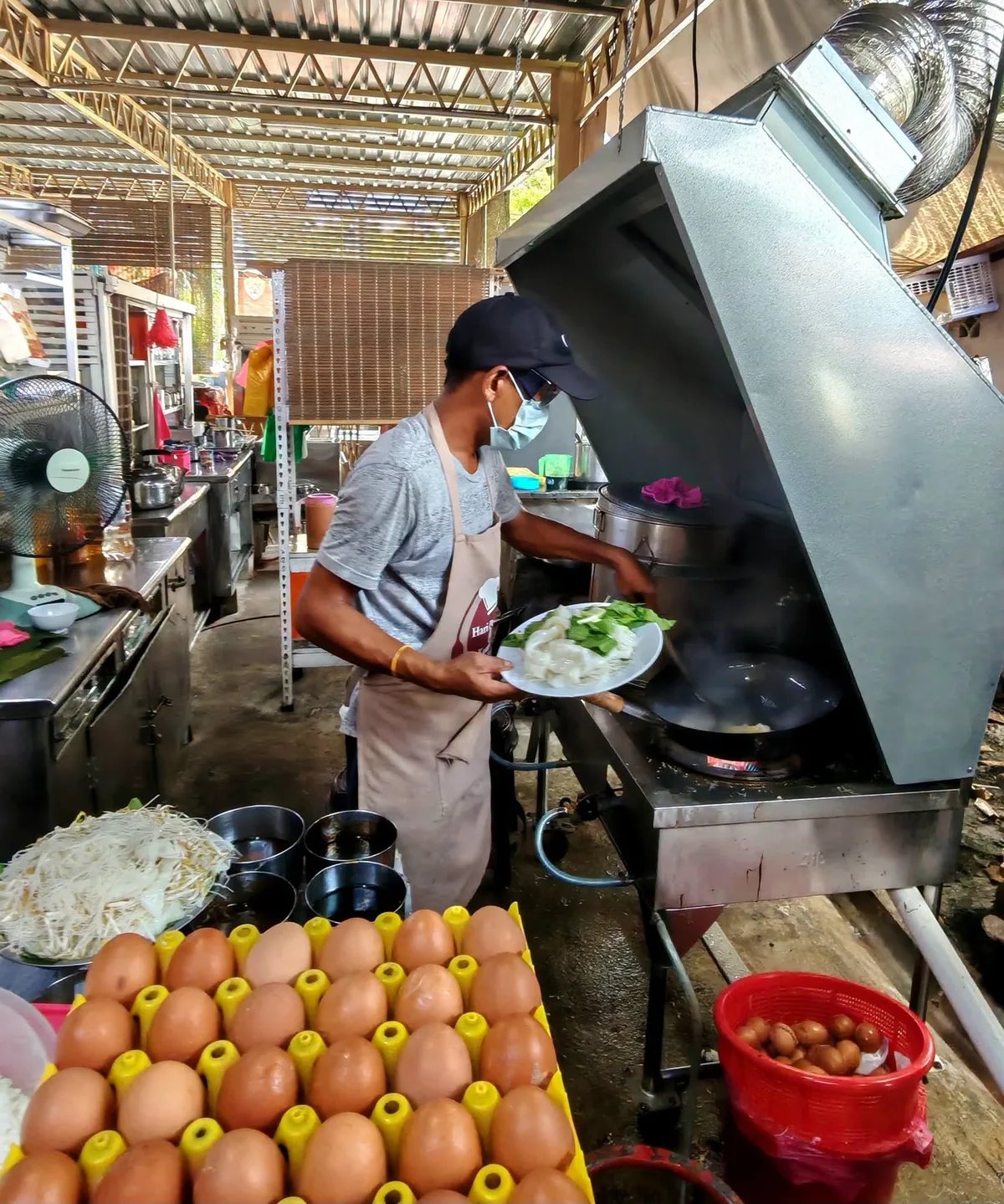 The stall hand tipping a plate of flat rice noodles and choy sum into the wok at Under The Tree, Lengkok Burma