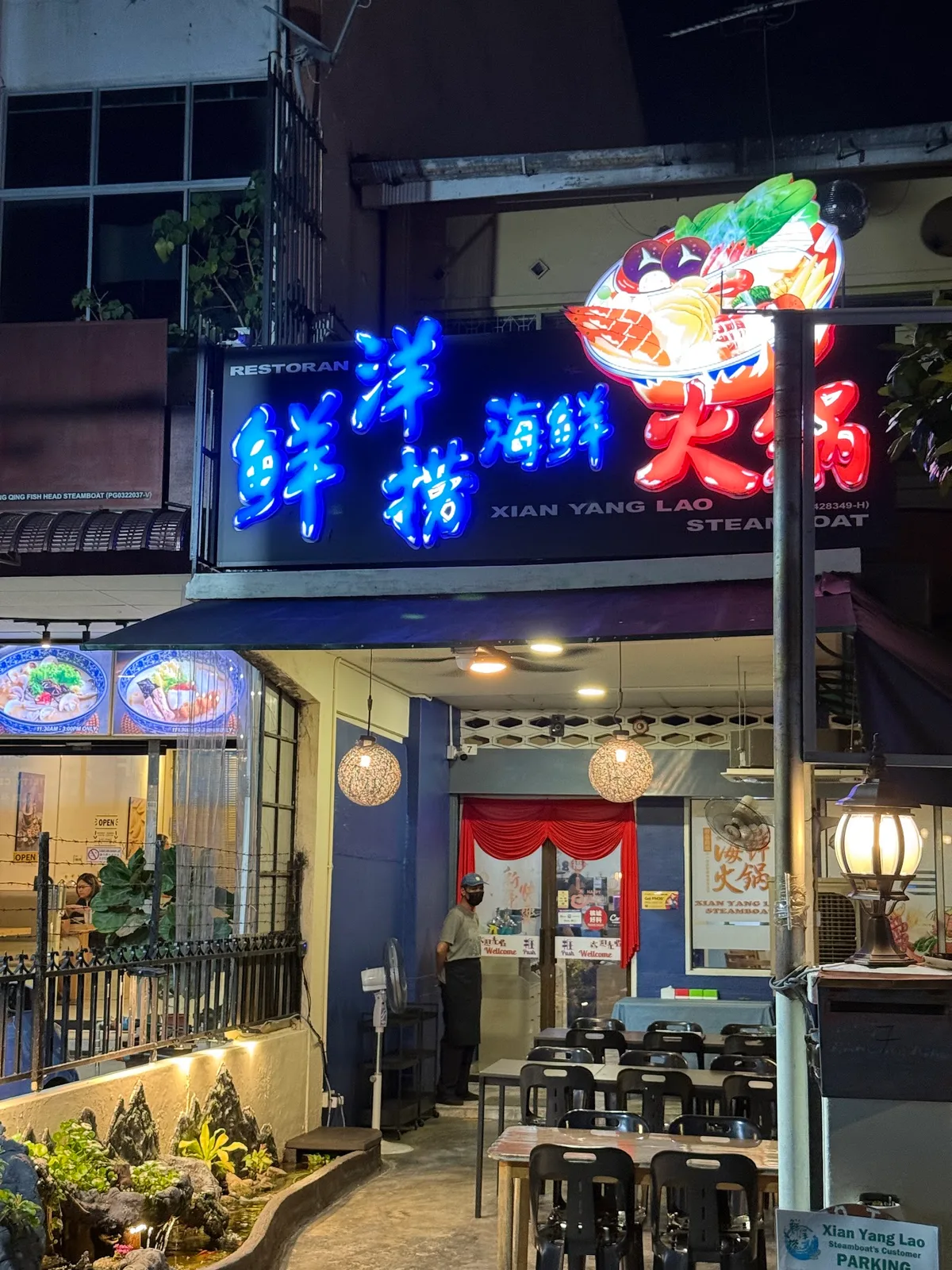 The restaurant frontage at night, with the lit Xian Yang Lao Steamboat signboard above the entrance and tables set up outside