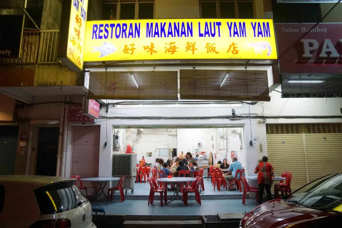 Lit yellow signboard reading Restoran Makanan Laut Yam Yam above the open shopfront, with diners at red plastic tables at night